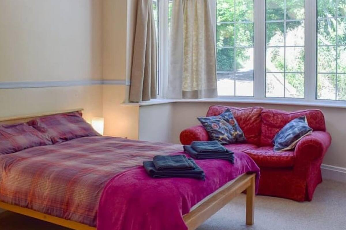 A bedroom with a bed, a bedside table, a red sofa, and a window with curtains.