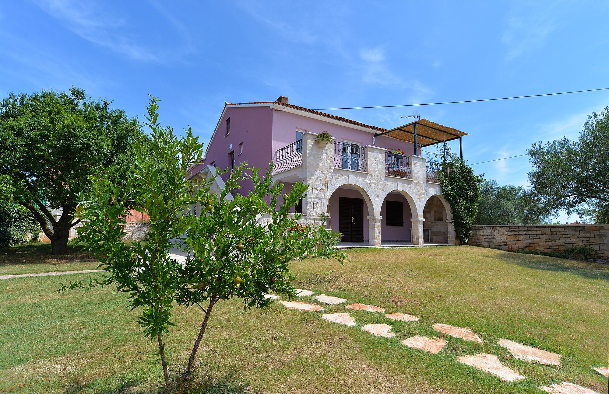 A two-story house with a pink facade, stone arches, and a balcony with a wooden pergola.