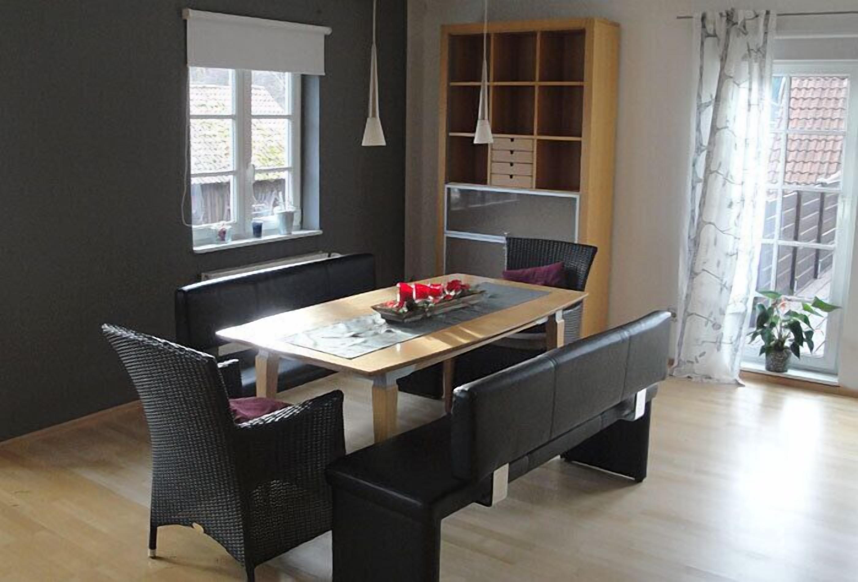 Un comedor con una mesa de madera, sillas negras y un banco. Hay una ventana con persianas, una estantería y una lámpara colgante.