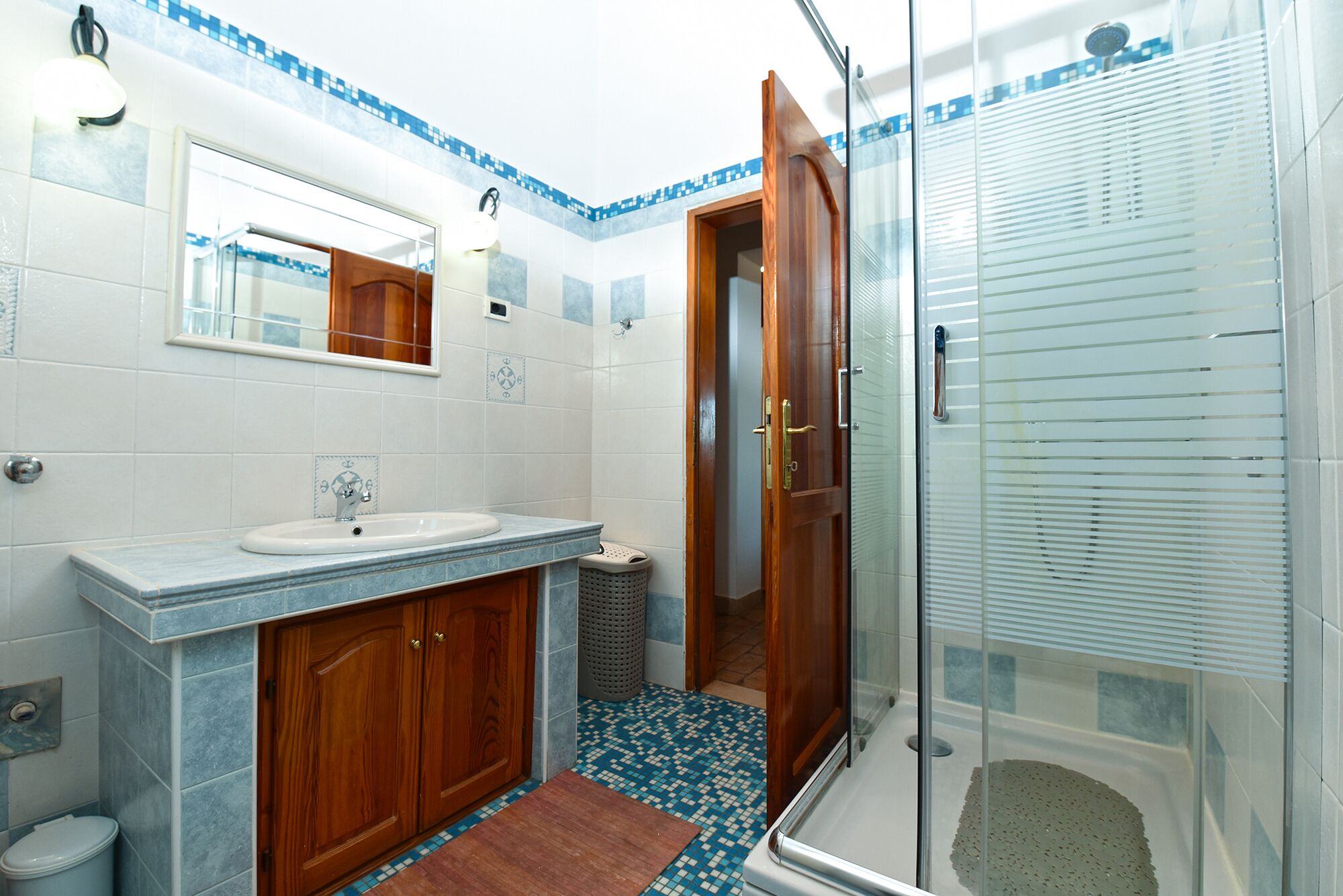 Een badkamer met een wastafel, spiegel, houten kastjes, een douche en een betegelde vloer.
