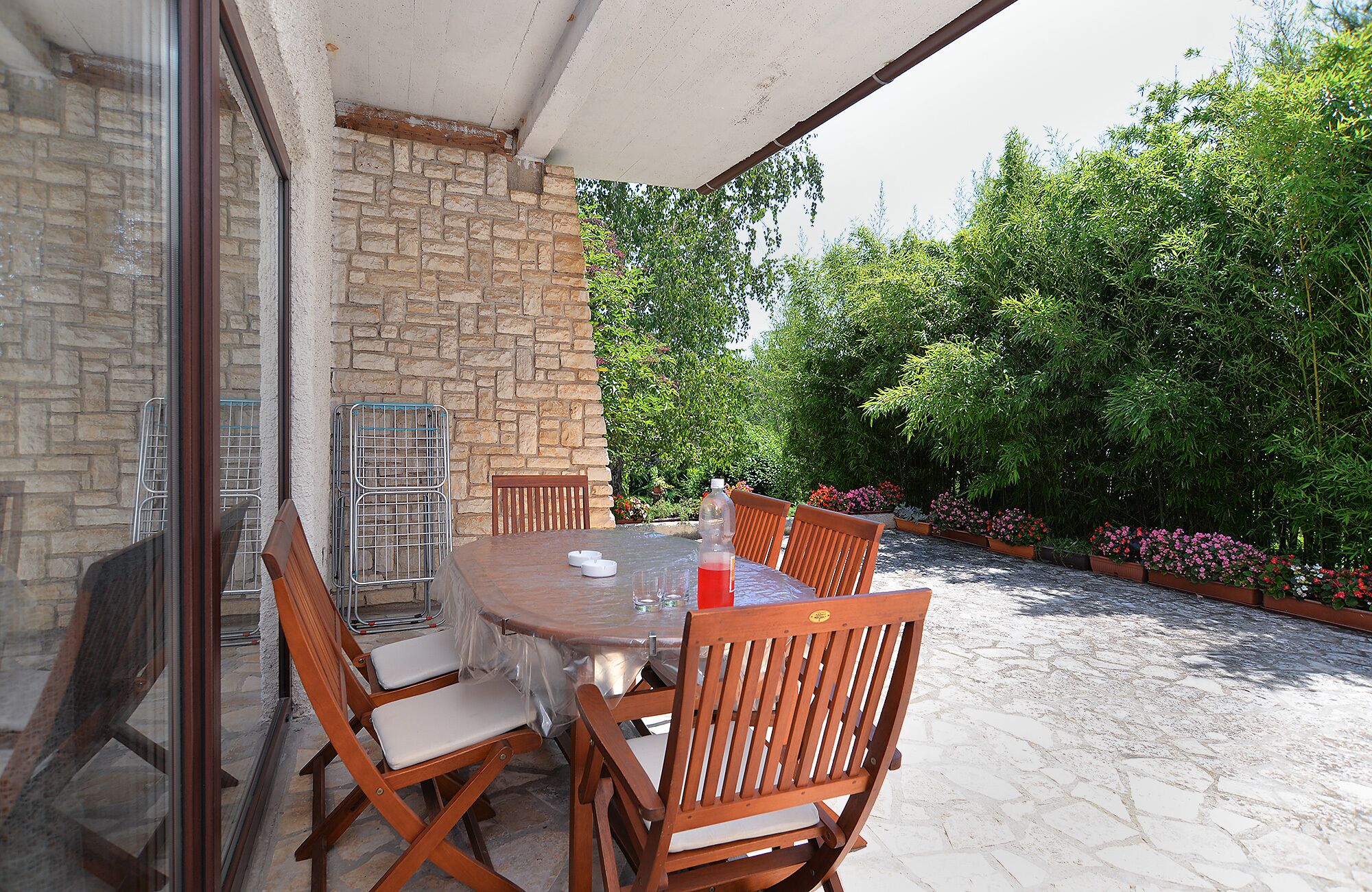 Un patio couvert avec un mur en pierre, une table ronde, des chaises en bois et une structure en verre qui l’entoure.