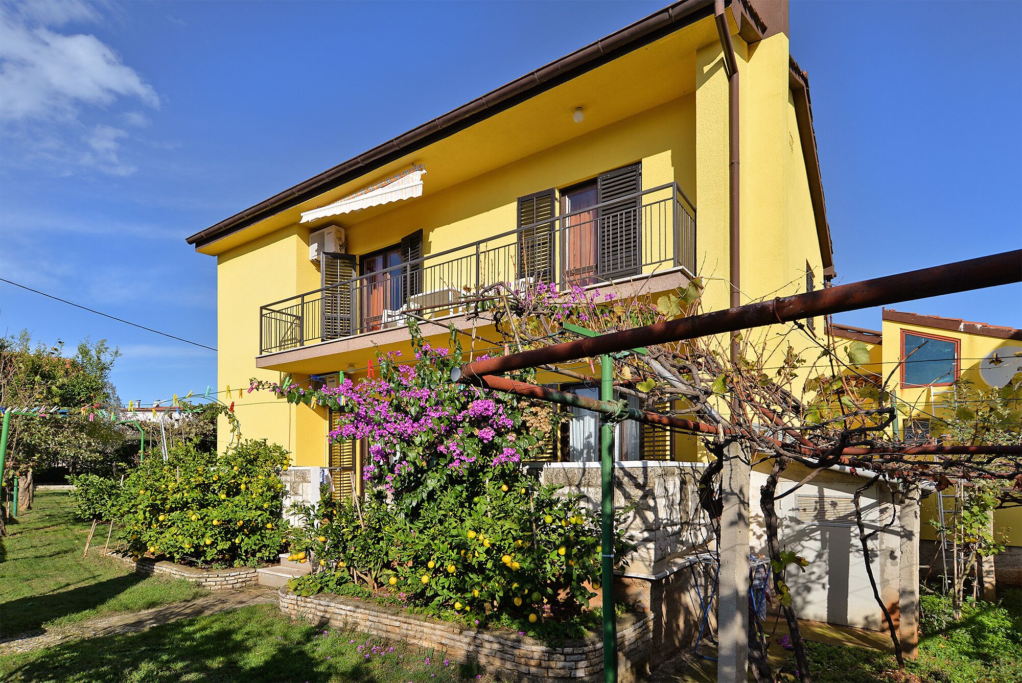 A yellow two-story house with a balcony, surrounded by a garden with various plants and flowers.