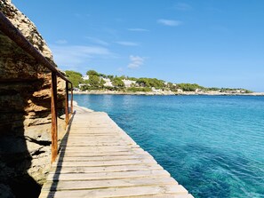 A wooden pier extending into clear blue water, with a rocky cliff on one side and a coastal village in the background. - Hotel Rocamarina - Adults Only (Santanyi)