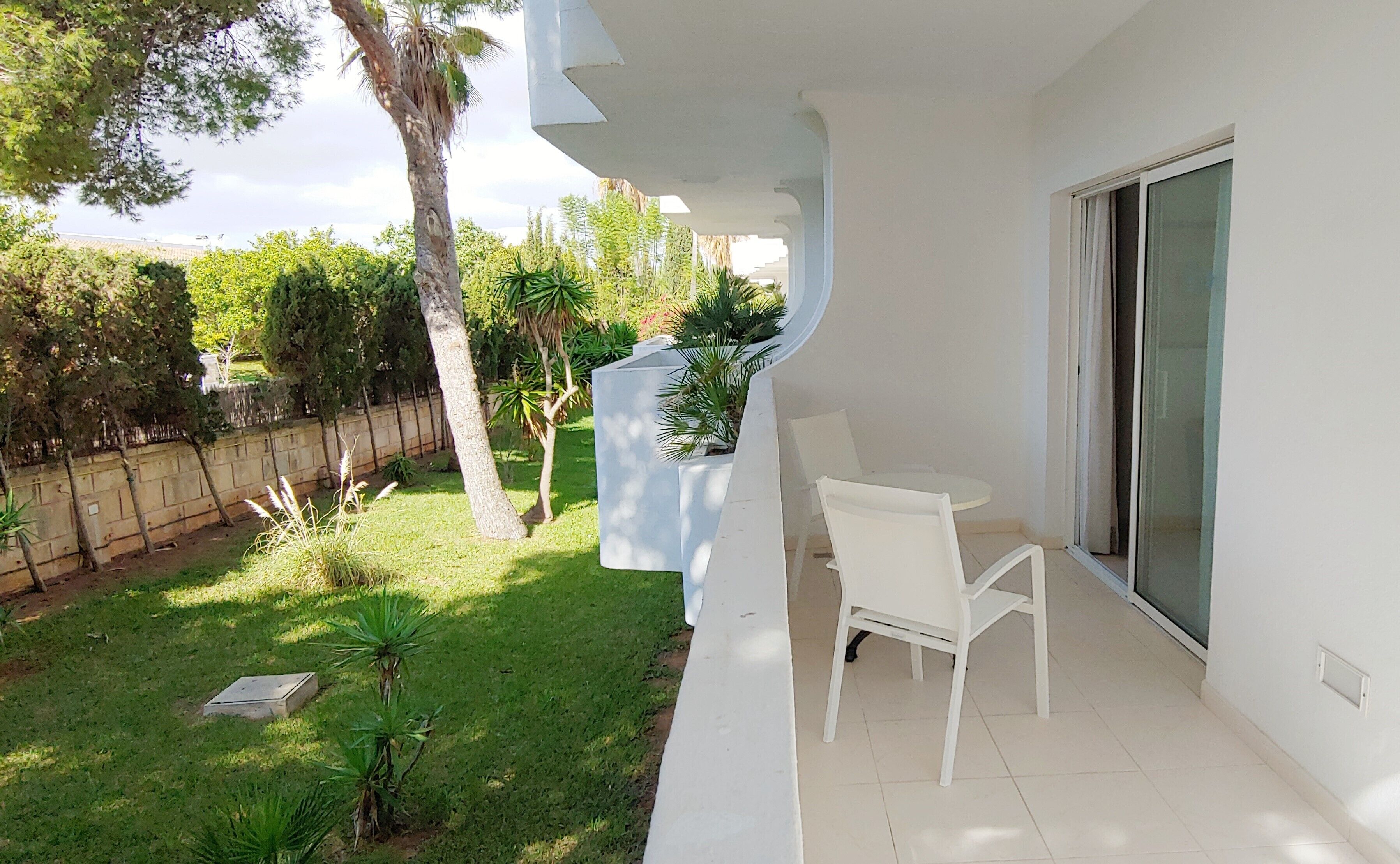 A balcony with a white table and chair, surrounded by greenery and a stone wall.
