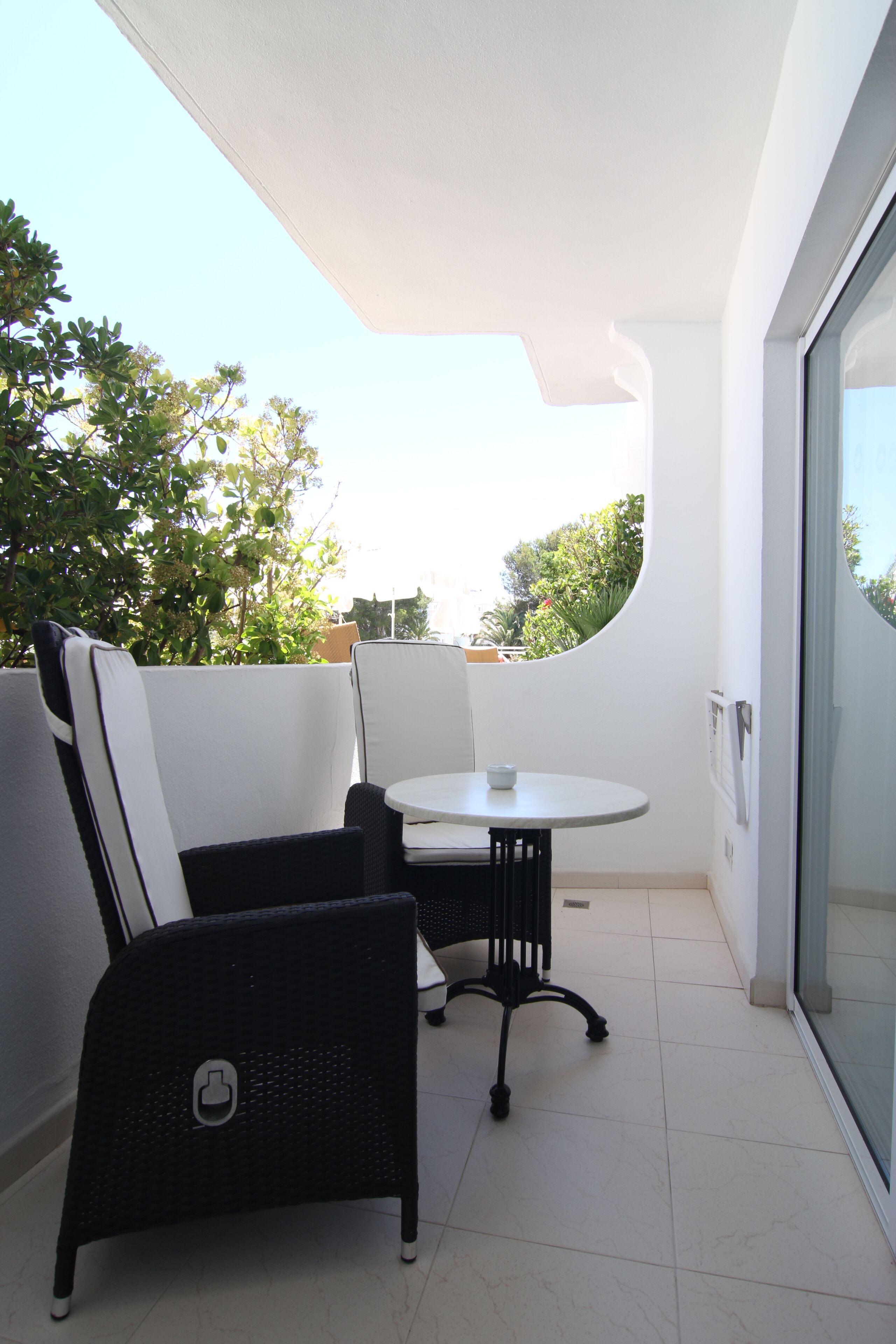 A balcony with a round table and two chairs, a glass door, and a view of trees and buildings.