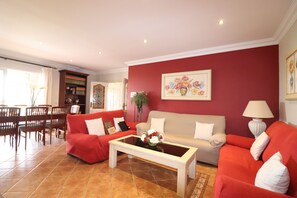 Een woonkamer met rode banken, een beige bank en een salontafel met bloemen. Er is een eethoek met houten stoelen en een tafel.