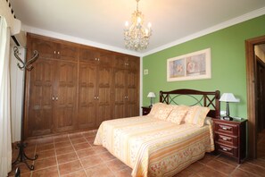 Een slaapkamer met een grote houten garderobe, een bed met bloemenbedekking, een kroonluchter en een groene muur met een ingelijst kunstwerk.