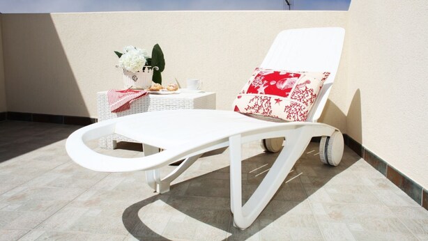 Una silla lounge blanca con un cojín de estampado rojo y blanco, una mesita con una taza blanca y un objeto decorativo, y una planta en maceta.