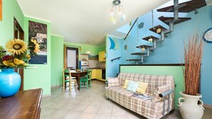 A living room with a plaid sofa, a dining area with a table and chairs, a kitchen in the background, and a staircase with hanging baskets. - I Tre Golfi Barbablù Apartment (San Vito Lo Capo)
