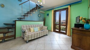 A living room with a sofa, a staircase, a TV, and a view through a glass door. - I Tre Golfi Barbablù Apartment (San Vito Lo Capo)