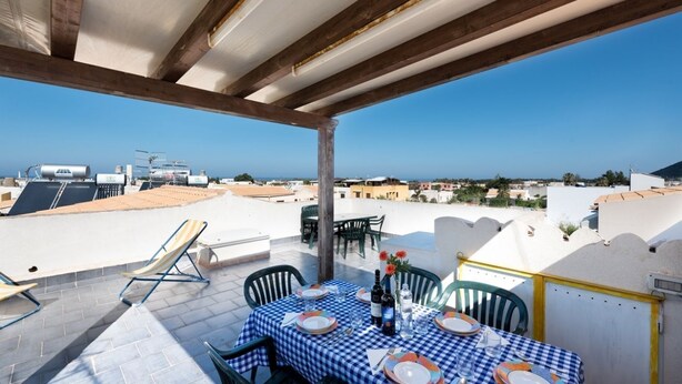 A rooftop terrace with a checkered tablecloth, outdoor chairs, and a view of the sea and buildings. - I Tre Golfi Barbablù Apartment (San Vito Lo Capo)