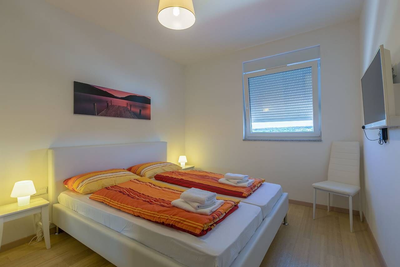 A bedroom with two beds, a desk, a chair, a TV, and a window with blinds.