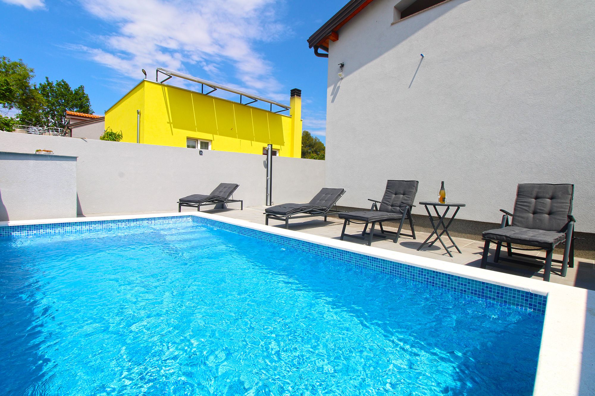 A swimming pool with lounge chairs and a table, surrounded by a white wall and a yellow building in the background.