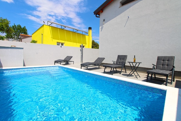 A swimming pool with lounge chairs and a table, surrounded by a white wall and a yellow building in the background.