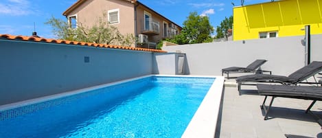 A swimming pool with lounge chairs, a tiled deck, and a building with a tiled roof.