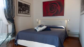 A bedroom with a bed, a bedside table, a glass cabinet, a framed photograph, and a large red rose painting.