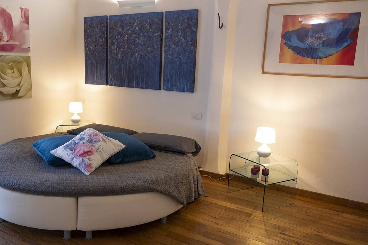 Ein rundes Bett mit grauer und weißer Bettwäsche, zwei Nachttischlampen und floralen Bildern an den Wänden.