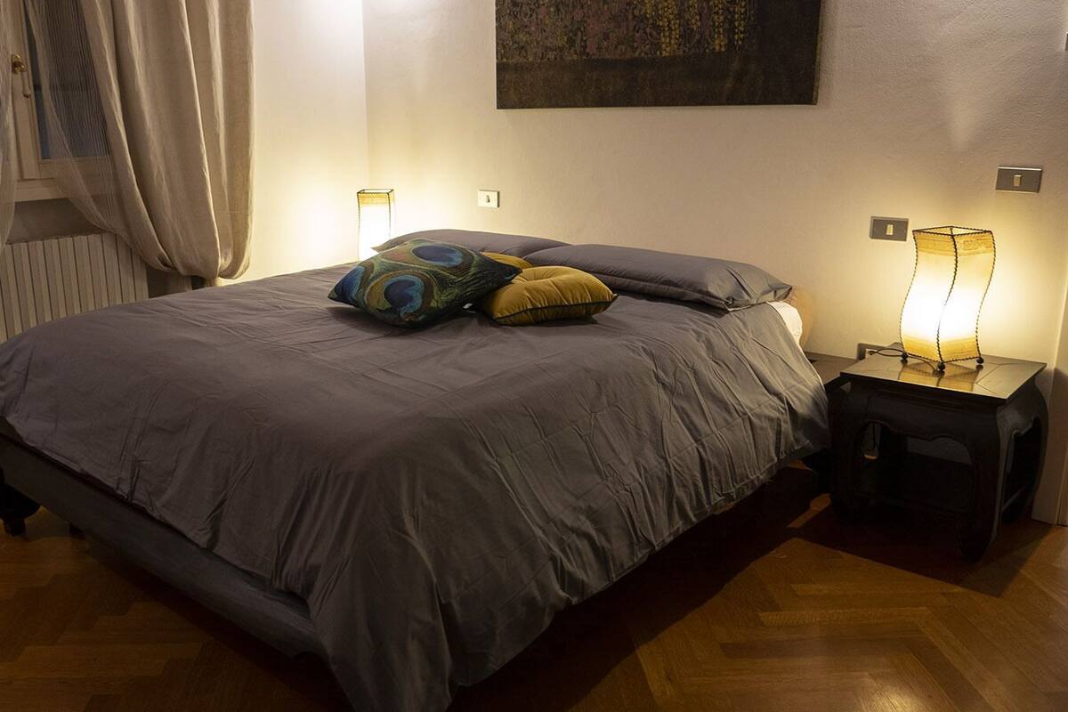 Ein ordentlich bezogenes Bett mit einer grauen Daunendecke, zwei Kissen und einem Nachttischchen mit einer Lampe.