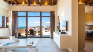 Un salon moderne doté d’une grande porte-fenêtre coulissante donnant sur un balcon avec vue sur la mer. Un téléviseur à écran plat est fixé au mur, ainsi qu’un canapé et une table basse sur laquelle se trouvent un livre et un objet décoratif.