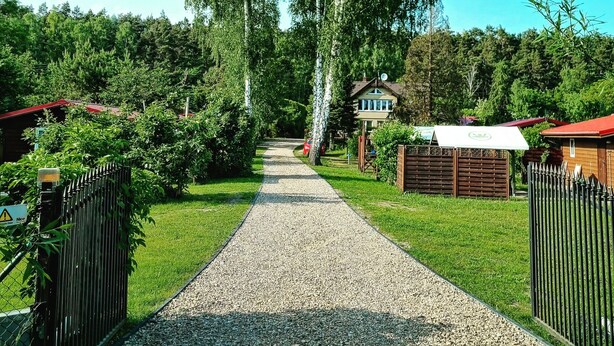 En grusgång leder fram till ett hus med rött tak, omgivet av frodig grönska och höga träd.