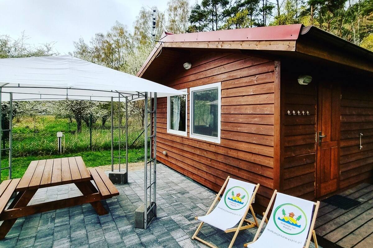 A wooden cabin with a covered patio area, a wooden table, and two lounge chairs with a logo.