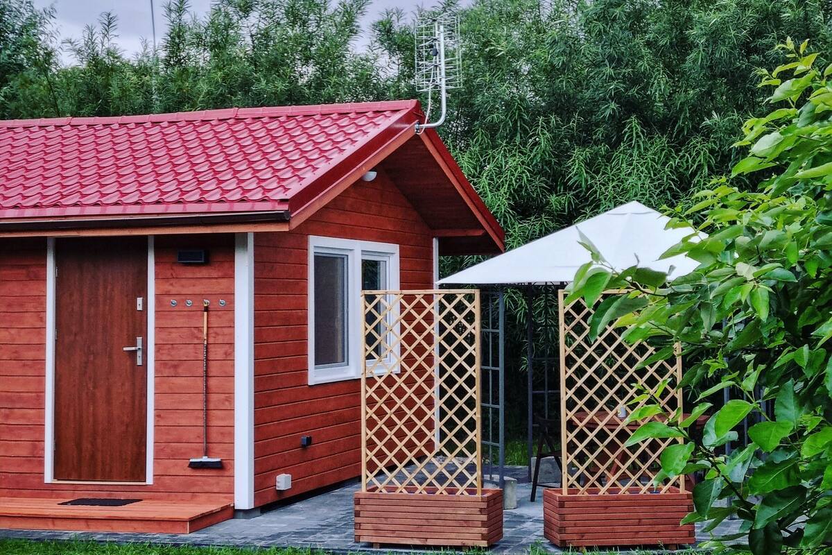 A small, red-roofed cabin with a wooden door and window, surrounded by a garden with a wooden fence and a white umbrella.