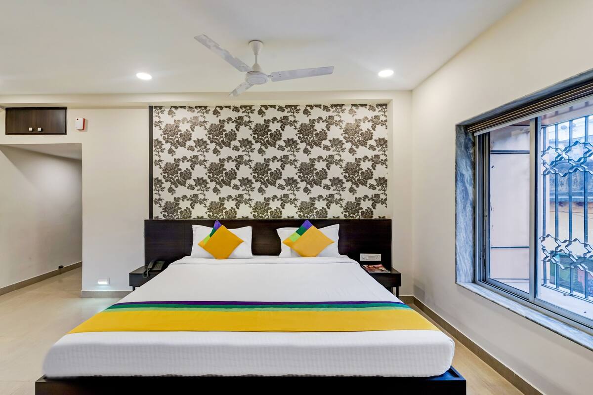 Une chambre d’hôtel avec un lit, un ventilateur de plafond, une fenêtre donnant sur un beau paysage et un mur orné d’un motif floral.
