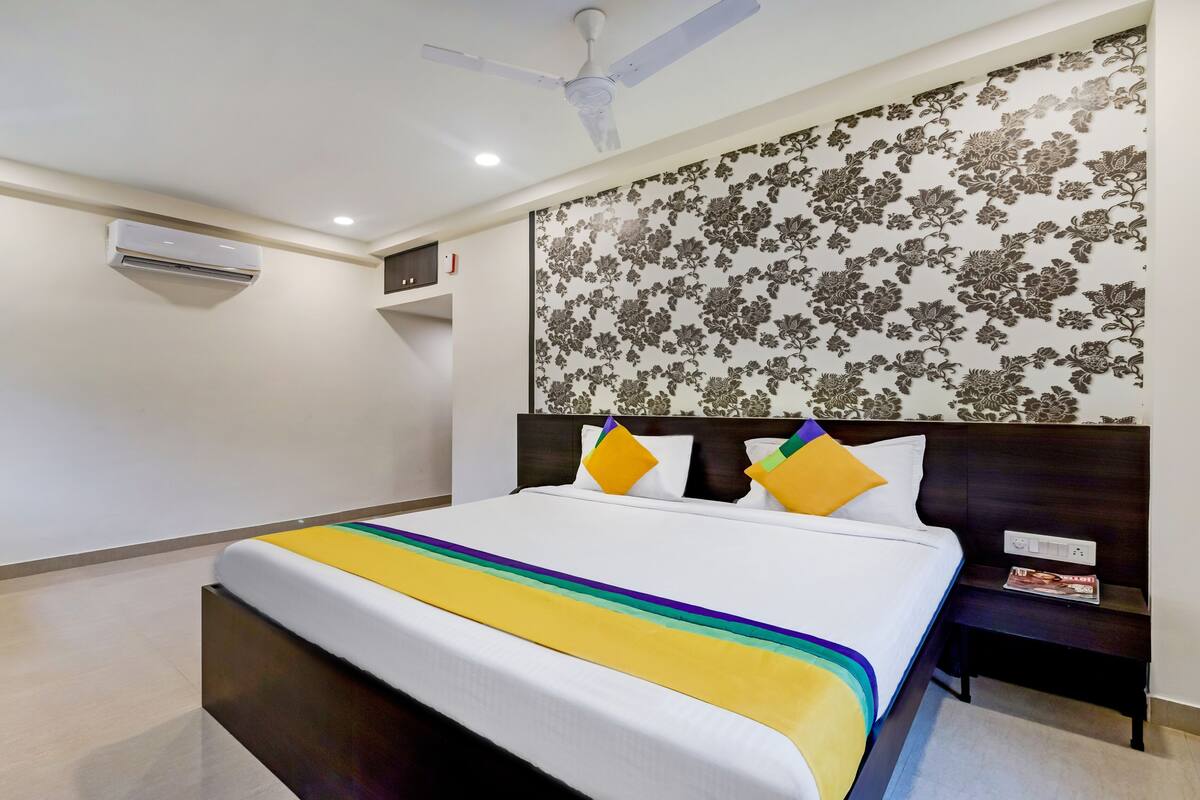 Une chambre d’hôtel comprenant un lit, une table de chevet, un climatiseur fixé au mur, un ventilateur de plafond et un papier peint à motifs floraux.
