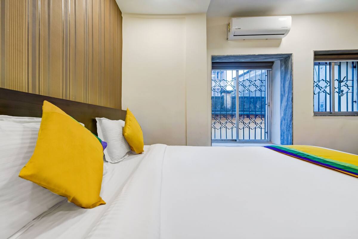 Une chambre d’hôtel avec un lit, des oreillers, un appareil de climatisation, une fenêtre avec des barreaux en métal et une vue sur l’extérieur.