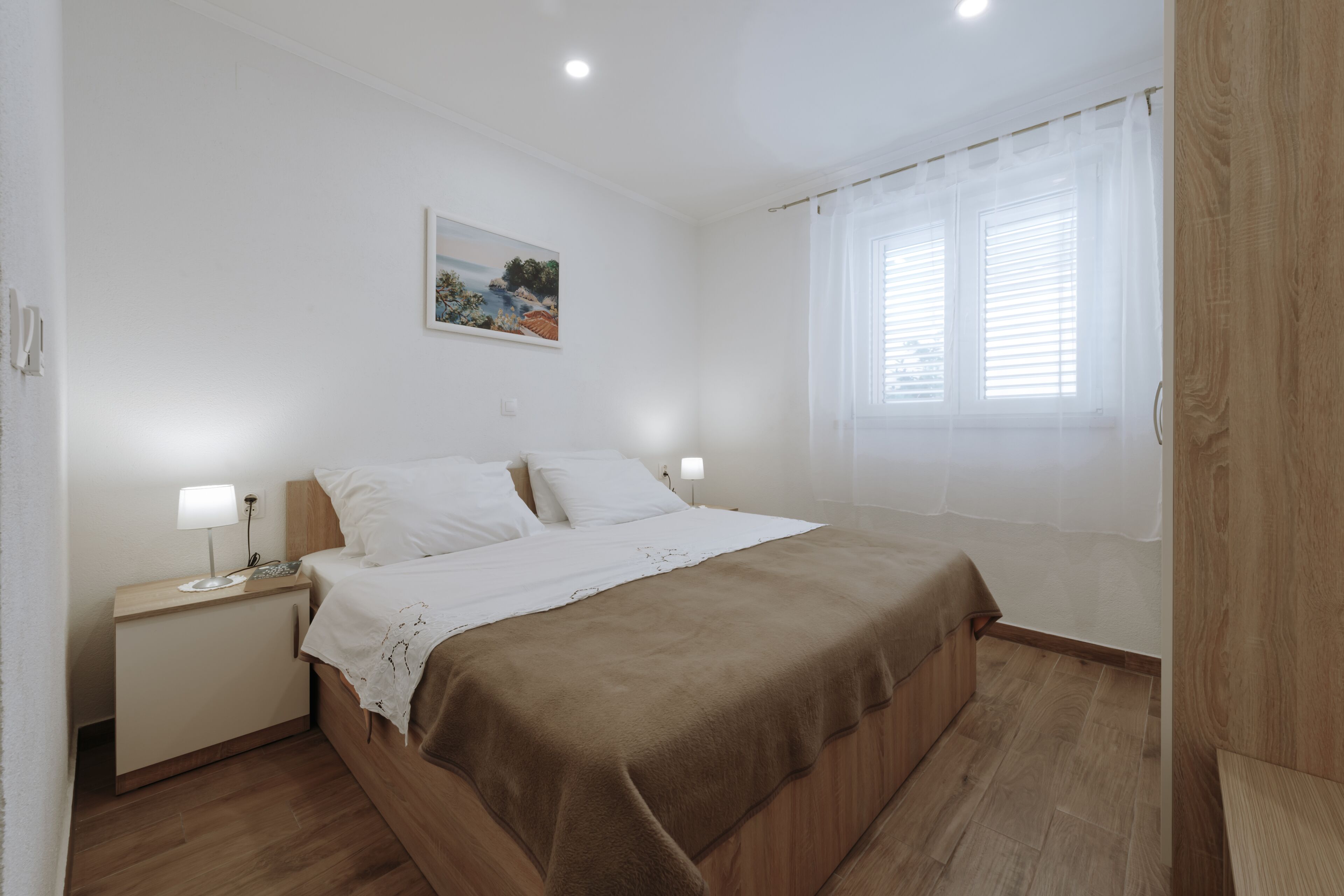 Un dormitorio con cama, mesitas de noche, un ventanal con cortinas y un cuadro colgado en la pared.