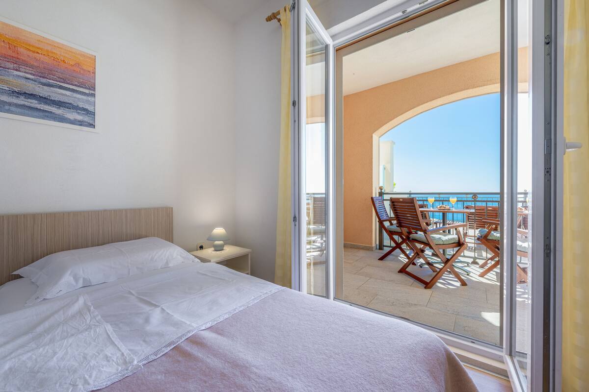 A hotel room with a bed, a nightstand, and a view of a balcony with outdoor furniture.