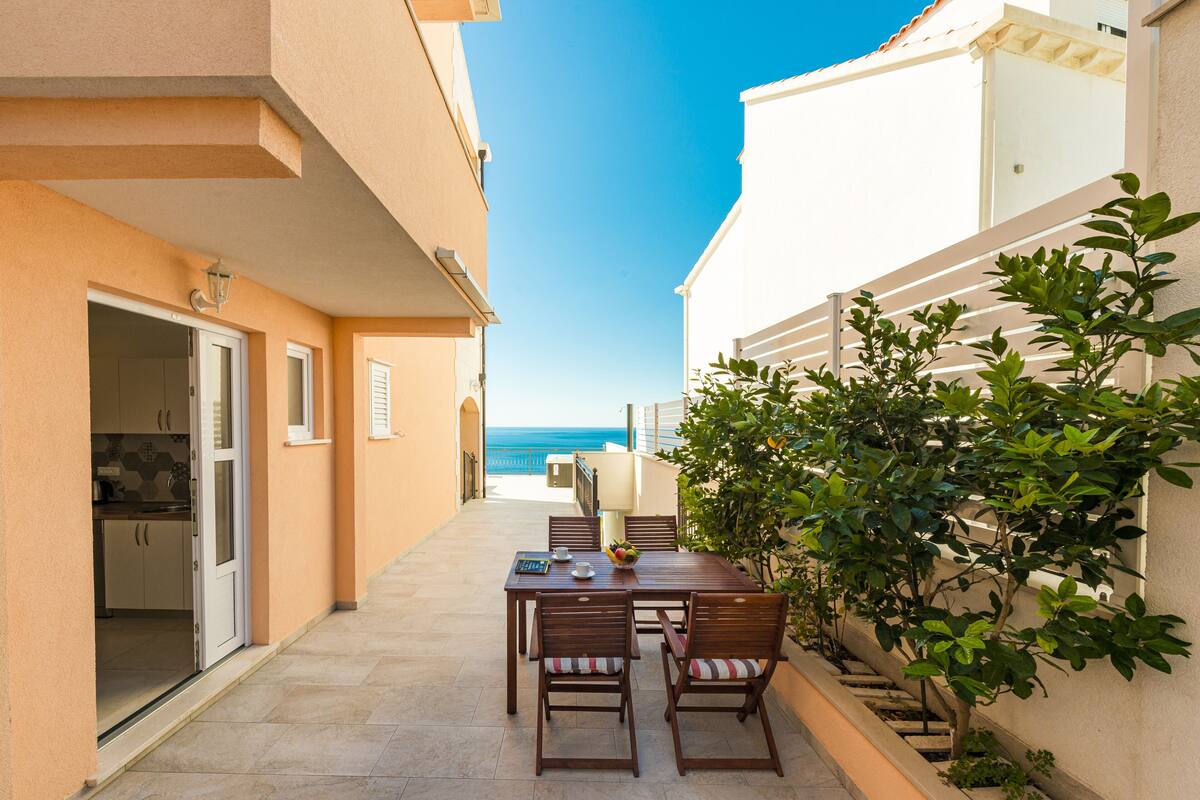 A narrow outdoor corridor with a dining area featuring a table and chairs, a small kitchen area, and a view of the sea.