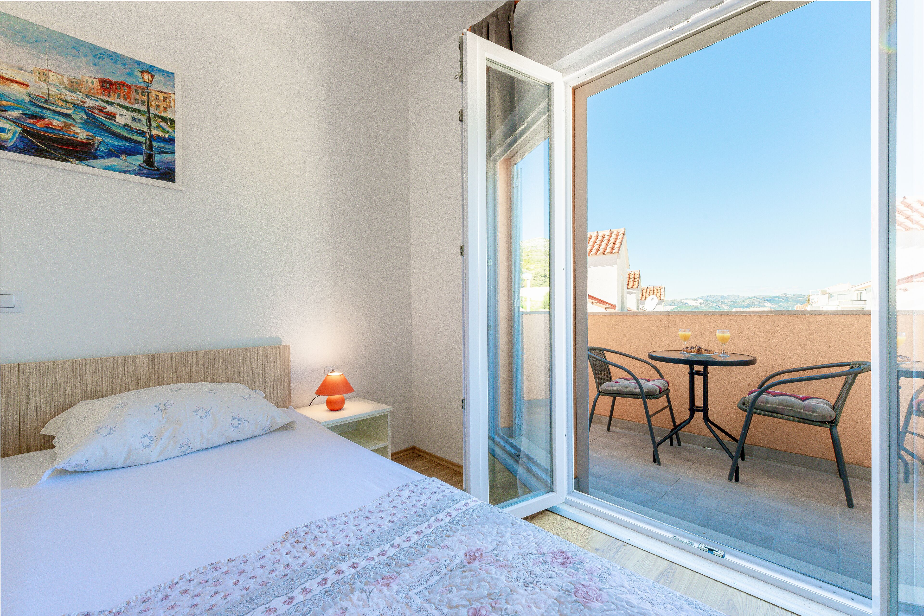 A hotel room with a bed, a nightstand with a lamp, a painting on the wall, and a balcony with chairs and a table.