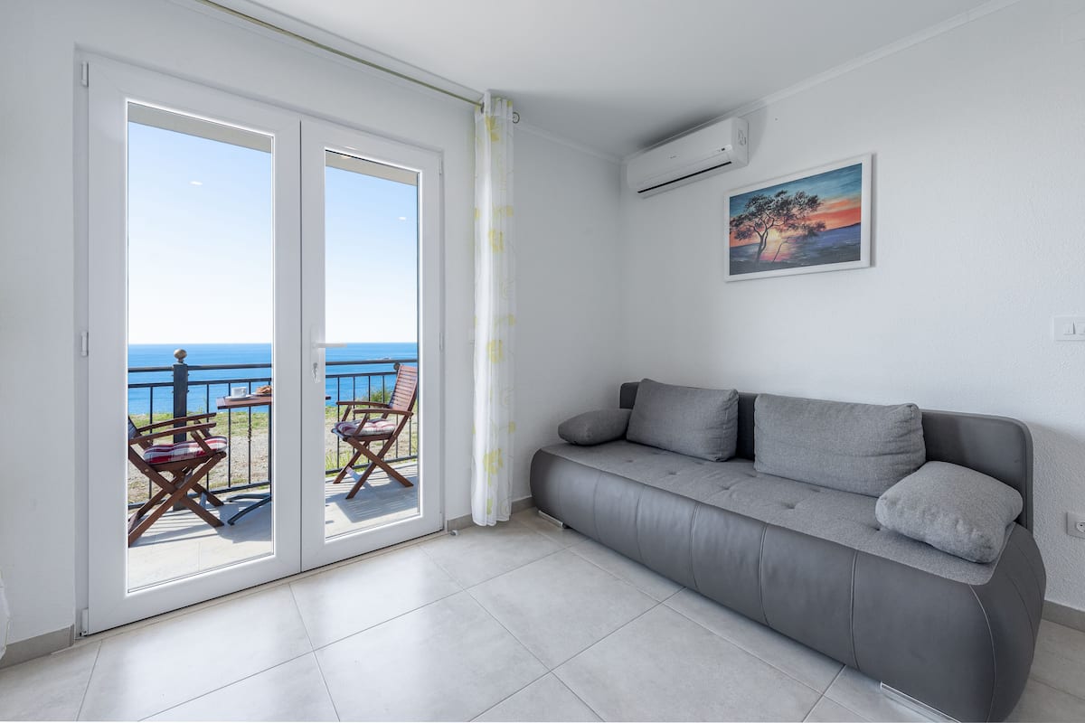 A modern living room with a grey sofa, a painting of a sunset, and a view of the sea through a glass door.