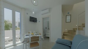 Ein modernes Wohnzimmer mit einer Couch, einem Esstisch mit Stühlen, einem an der Wand montierten Fernseher und einer Treppe, die zu einer oberen Etage führt.