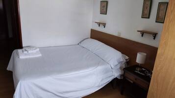Una cama bien tendida con sábanas blancas, cabecera de madera y mesita de noche con lámpara.