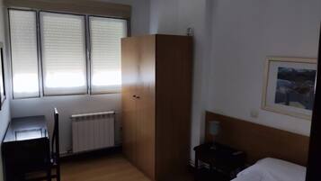 A hotel room with a bed, a desk, a radiator, a wardrobe, and a window with blinds.