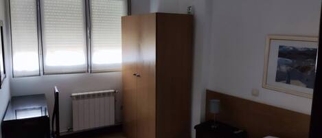 A hotel room with a bed, a desk, a radiator, a wardrobe, and a window with blinds.