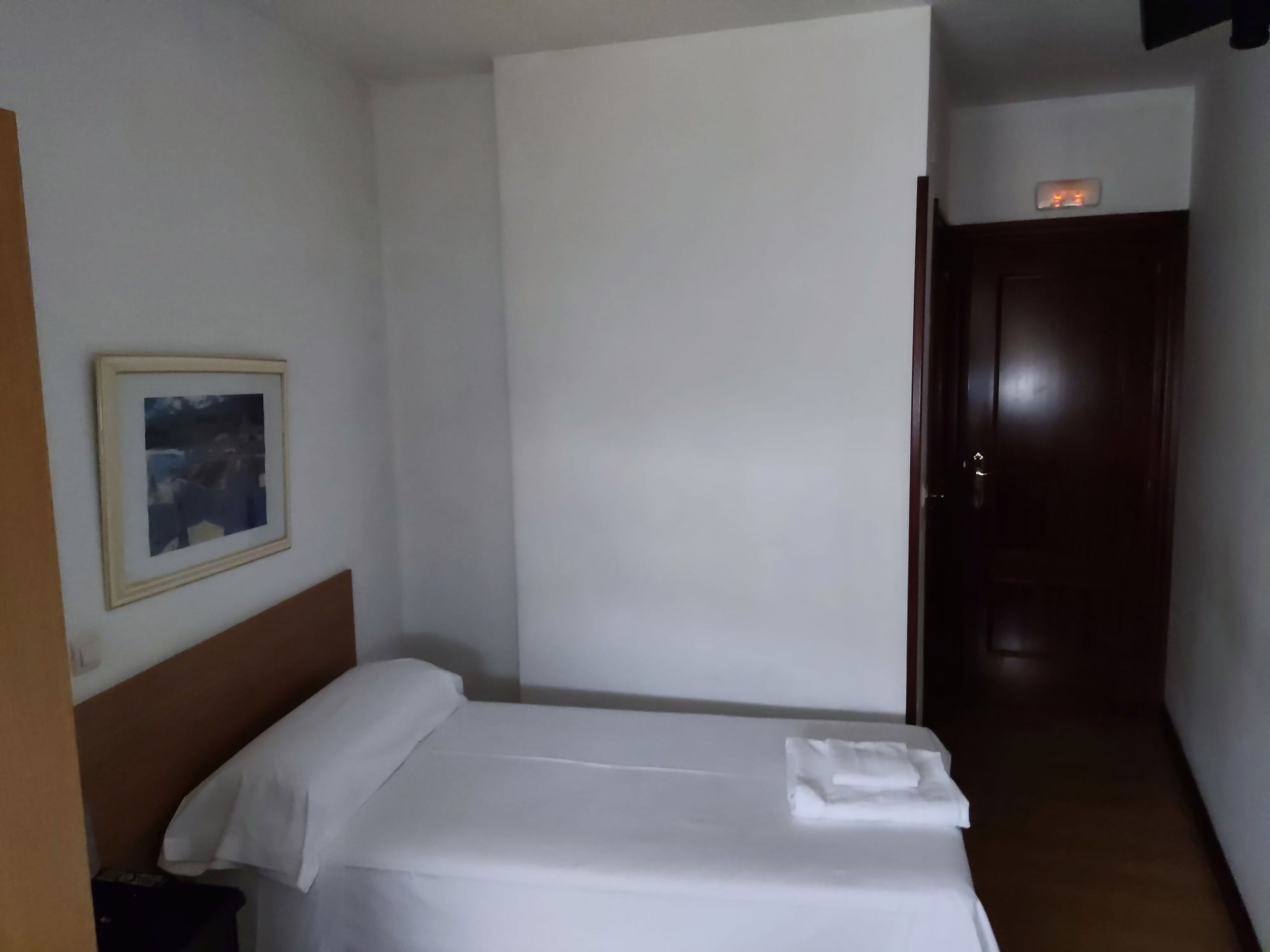 Ett hotellrum med en säng, en inramad tavla på väggen och en dörr som leder till ett annat rum.