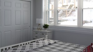 A bedroom with a white bed, a nightstand with a lamp, a window with blinds, and a door.