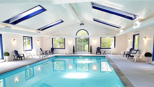 An indoor swimming pool with clear blue water, surrounded by lounge chairs and tables, under a high ceiling with skylights.