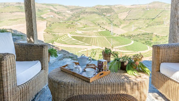 Un set di seduta e tavolino in vimini con un vassoio contenente tè, tazze e pane, con vista su vigneti a terrazza.