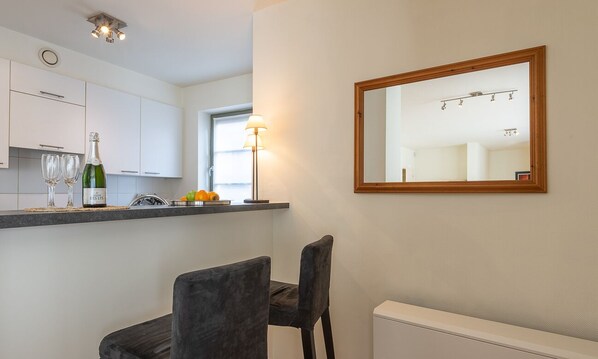 A modern kitchen with white cabinets, a countertop with a bottle of champagne, glasses, and a fruit bowl. There is a chair and a mirror on the wall.