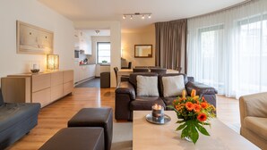 A modern living room with a sofa, ottoman, and a table with a candle and flowers. There is a kitchen in the background with cabinets and a dining area.