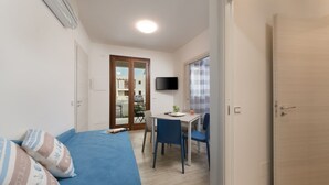 Une pièce de taille modeste, avec un canapé bleu, une petite table à manger entourée de chaises, un téléviseur fixé au mur et une porte donnant sur une autre pièce.