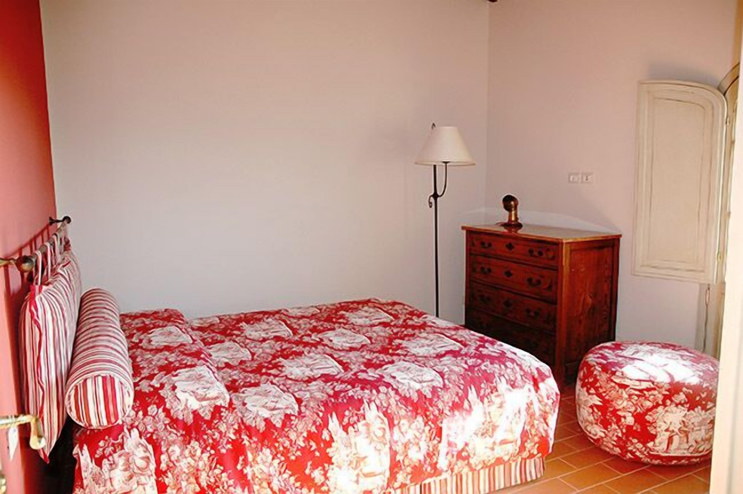 A bedroom with a floral bedspread, a nightstand, a lamp, a dresser, and a door.