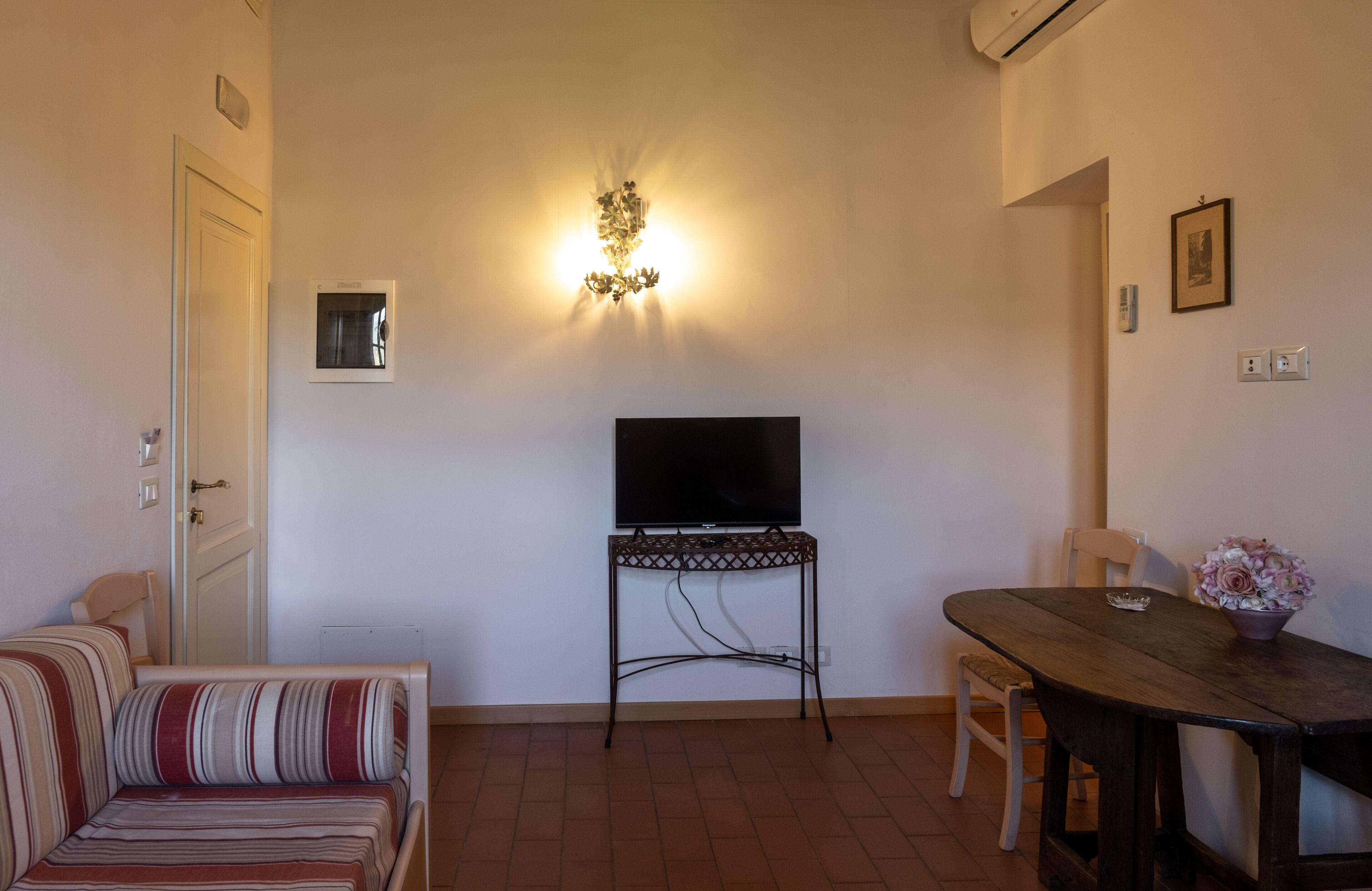 Una habitación pequeña y acogedora con sofá, mesa de comedor, televisor sobre un mueble bajo y una lámpara fijada a la pared.