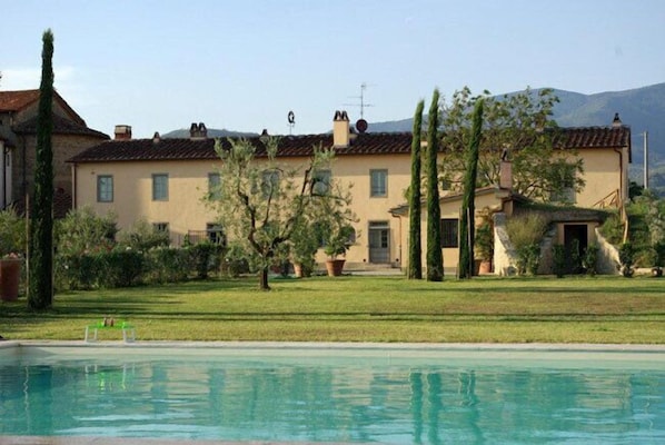 A poolside view of a building with a tiled roof, surrounded by greenery and a clear sky.