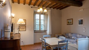 Ein gemütliches Zimmer mit Holzbalkendecke, einem Esstisch mit Stühlen, einer Couch und einem Fenster mit Vorhängen.