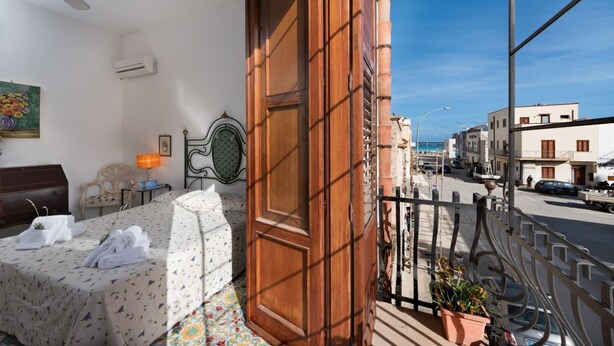 Ein Balkon mit Holztür, einem Bett mit Blumenmuster-Bettdecke, einem kleinen Tisch mit Lampe und Blick auf die Straße sowie die gegenüberliegenden Gebäude.